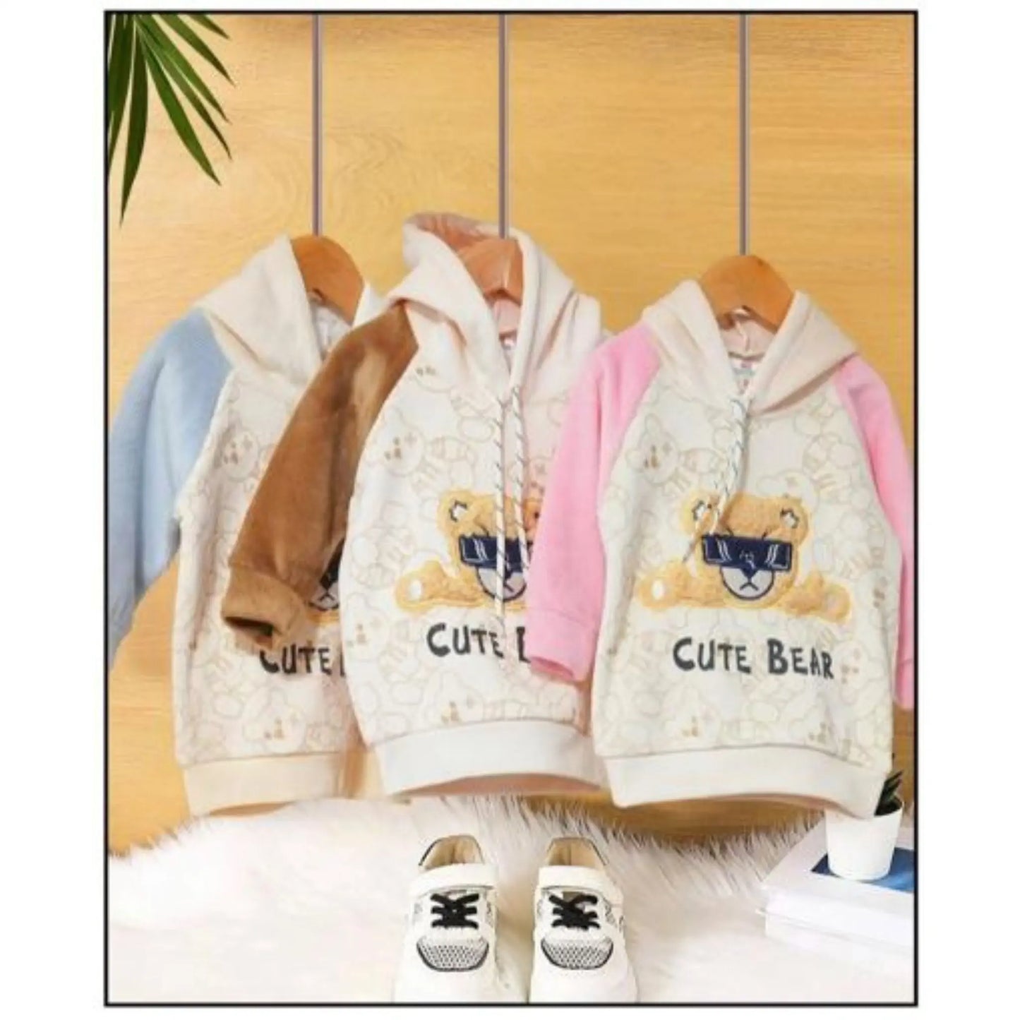 Children's hoodies with teddy bear design and 'CUTE BEAR' text on hangers against a wooden panel background.