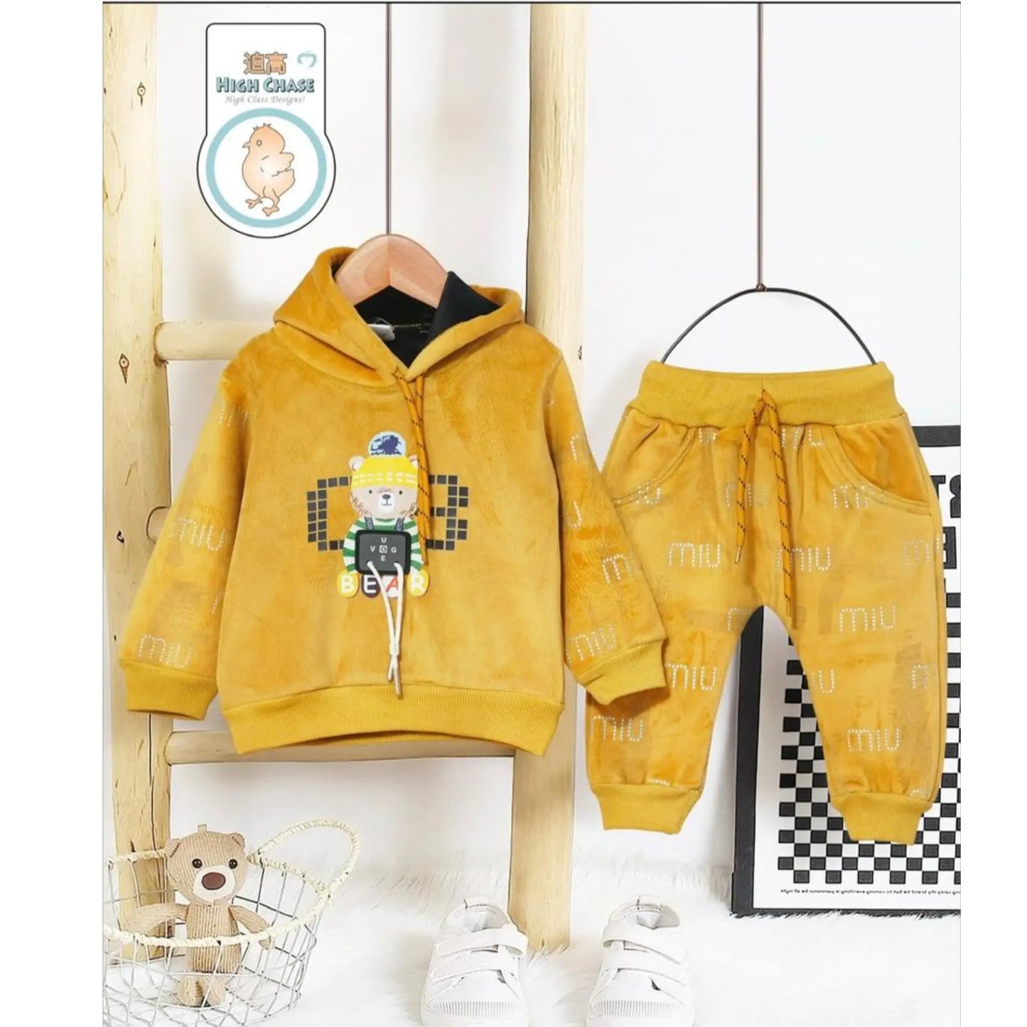 Yellow children's hoodie and pants set on a wooden ladder with a teddy bear and checkered pattern in the background.