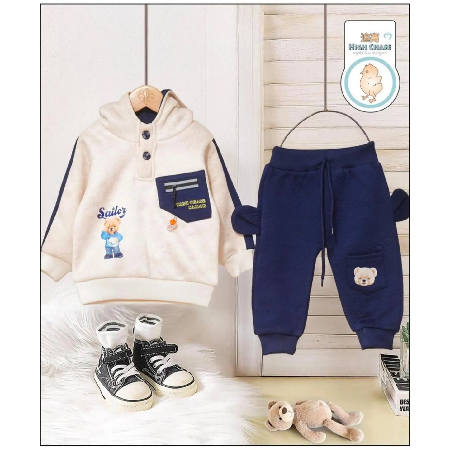 Children's clothing set with hoodie, pants, shoes, and teddy bear plush toy on a wooden floor.